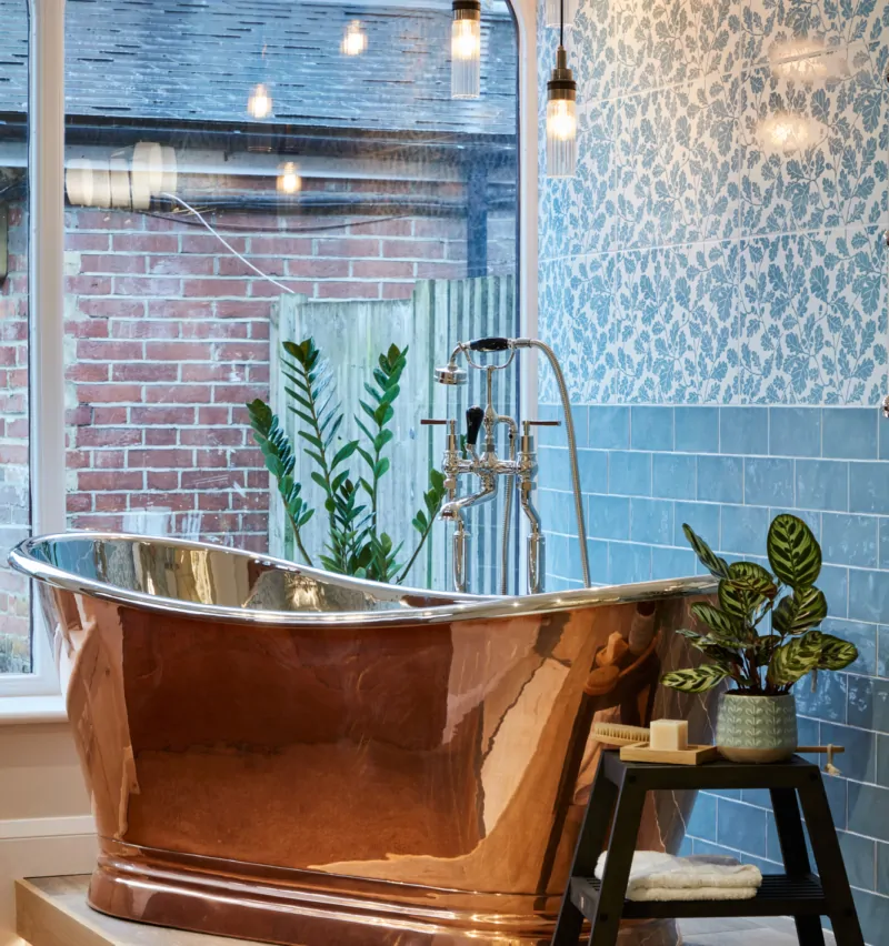Copper bathtub beside plants, with patterned wallpaper and wooden flooring in a bright bathroom.