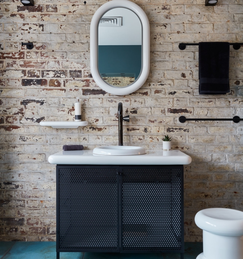 Bathroom with an oval mirror, exposed brick wall, and turquoise tiled floor.