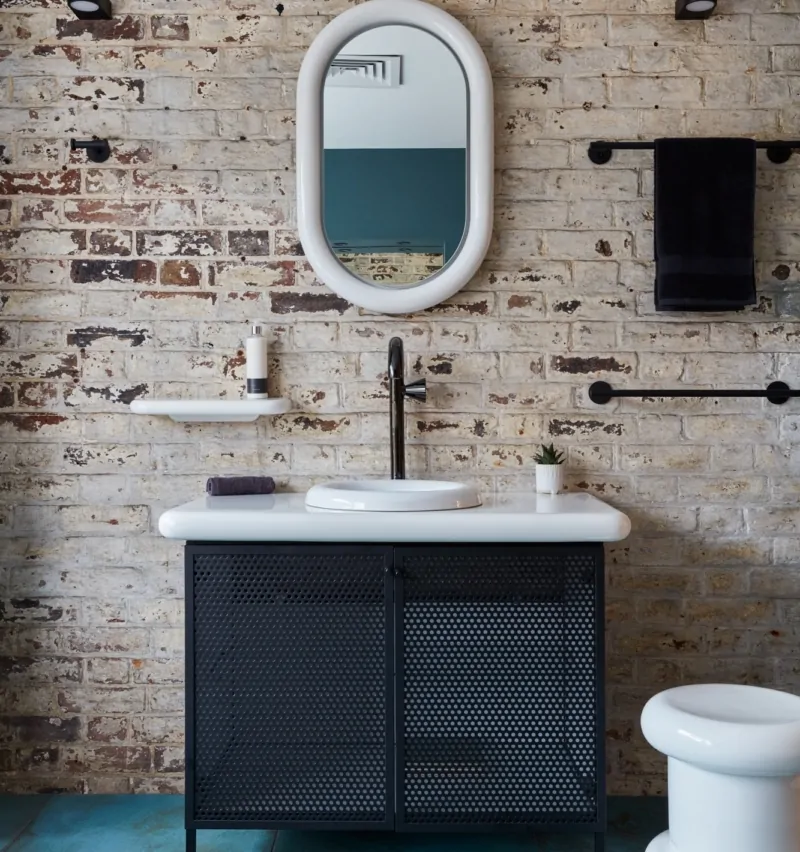Bathroom with an oval mirror, exposed brick wall, and turquoise tiled floor.