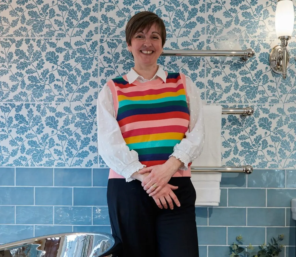 Alison standing in a bathroom display with blue floral wall tiles, a chrome towel rail, and a freestanding copper bathtub