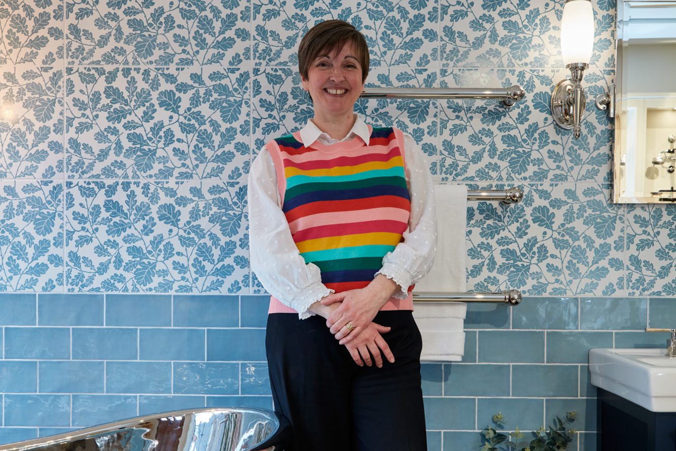 Alison standing in a bathroom display with blue floral wall tiles, a chrome towel rail, and a freestanding copper bathtub