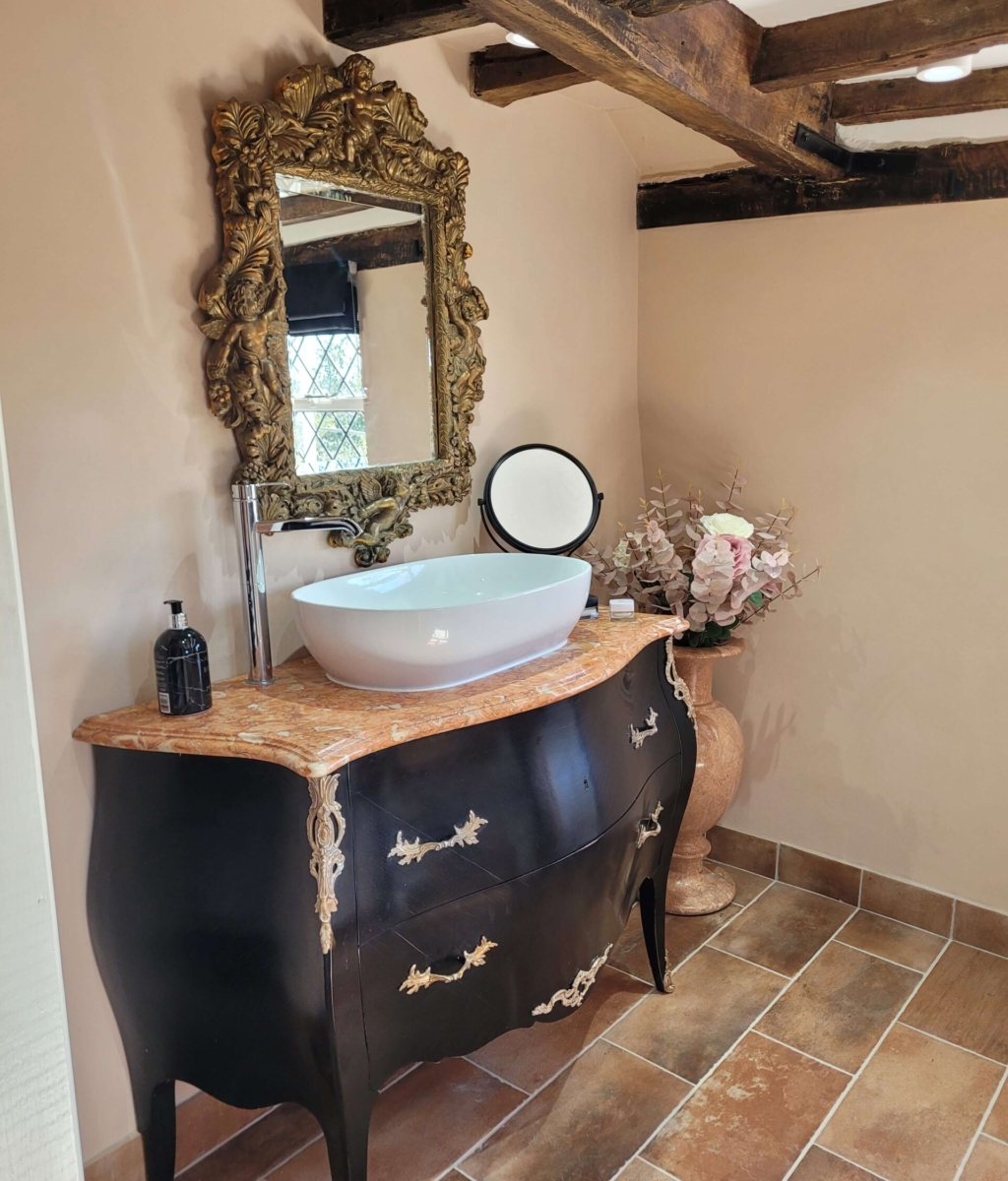 Antique-style bathroom with a gold-framed ornate mirror, black vanity unit with brass details, marble countertop, vessel sink, and exposed wooden beams