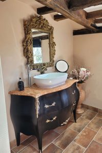 Antique-style bathroom with a gold-framed ornate mirror, black vanity unit with brass details, marble countertop, vessel sink, and exposed wooden beams