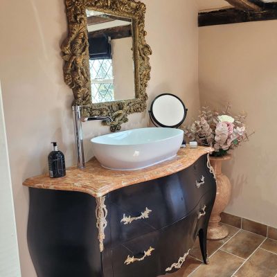 Antique-style bathroom with a gold-framed ornate mirror, black vanity unit with brass details, marble countertop, vessel sink, and exposed wooden beams