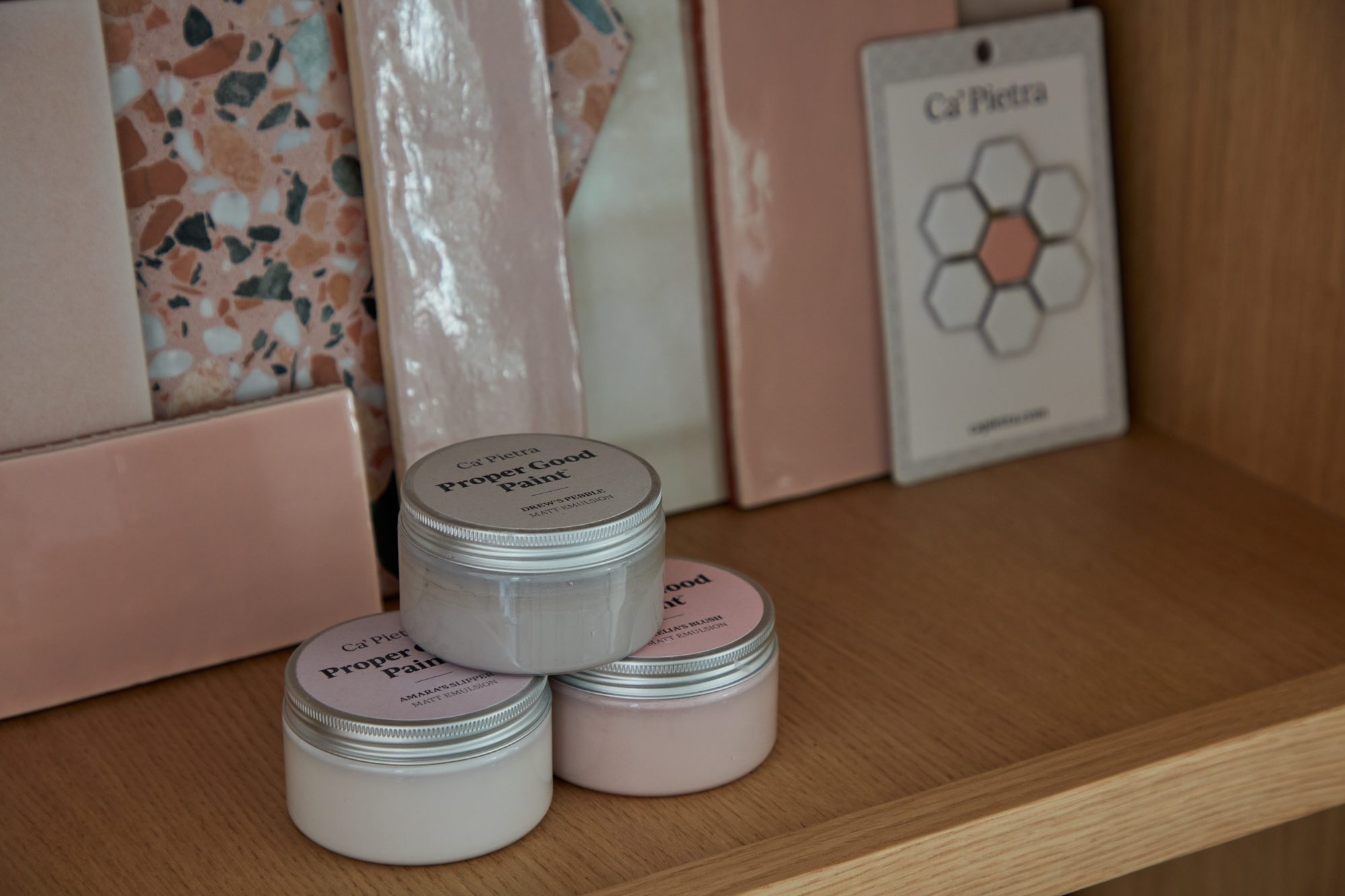 Samples of pink terrazzo tiles and pastel-colored paint jars from Ca’ Pietra on a wooden shelf