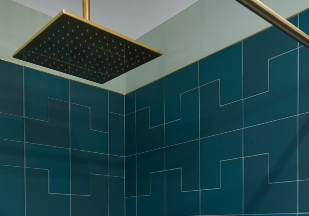Square showerhead with brass finish over teal geometric tiled shower walls.
