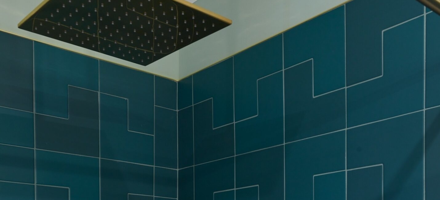 Square showerhead with brass finish over teal geometric tiled shower walls.
