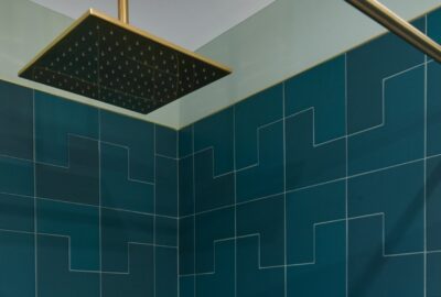 Square showerhead with brass finish over teal geometric tiled shower walls.