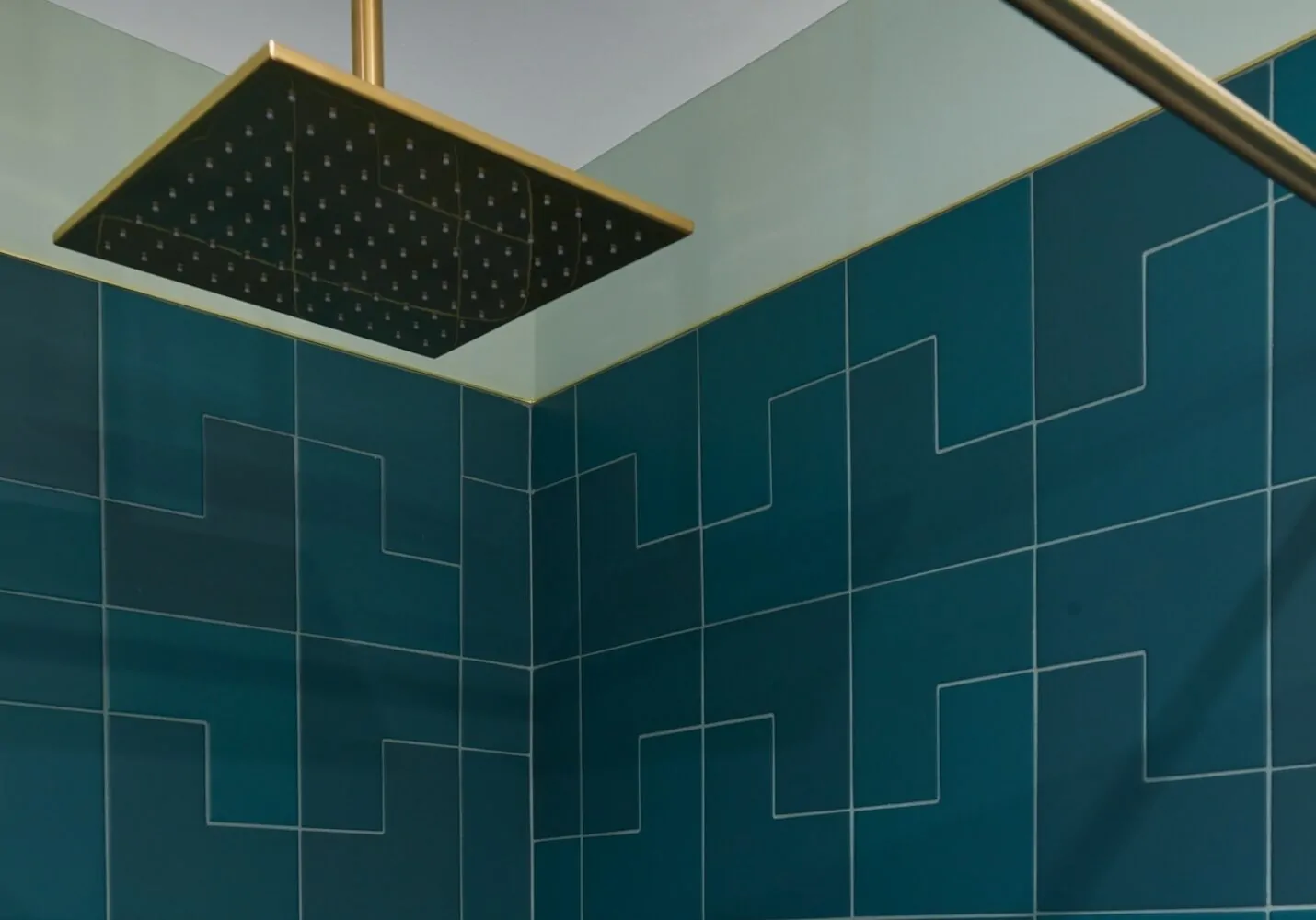 Square showerhead with brass finish over teal geometric tiled shower walls.