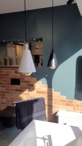 Bathroom with black and white pendant lights, black towel rail with towel, exposed brick wall, and freestanding bathtub corner