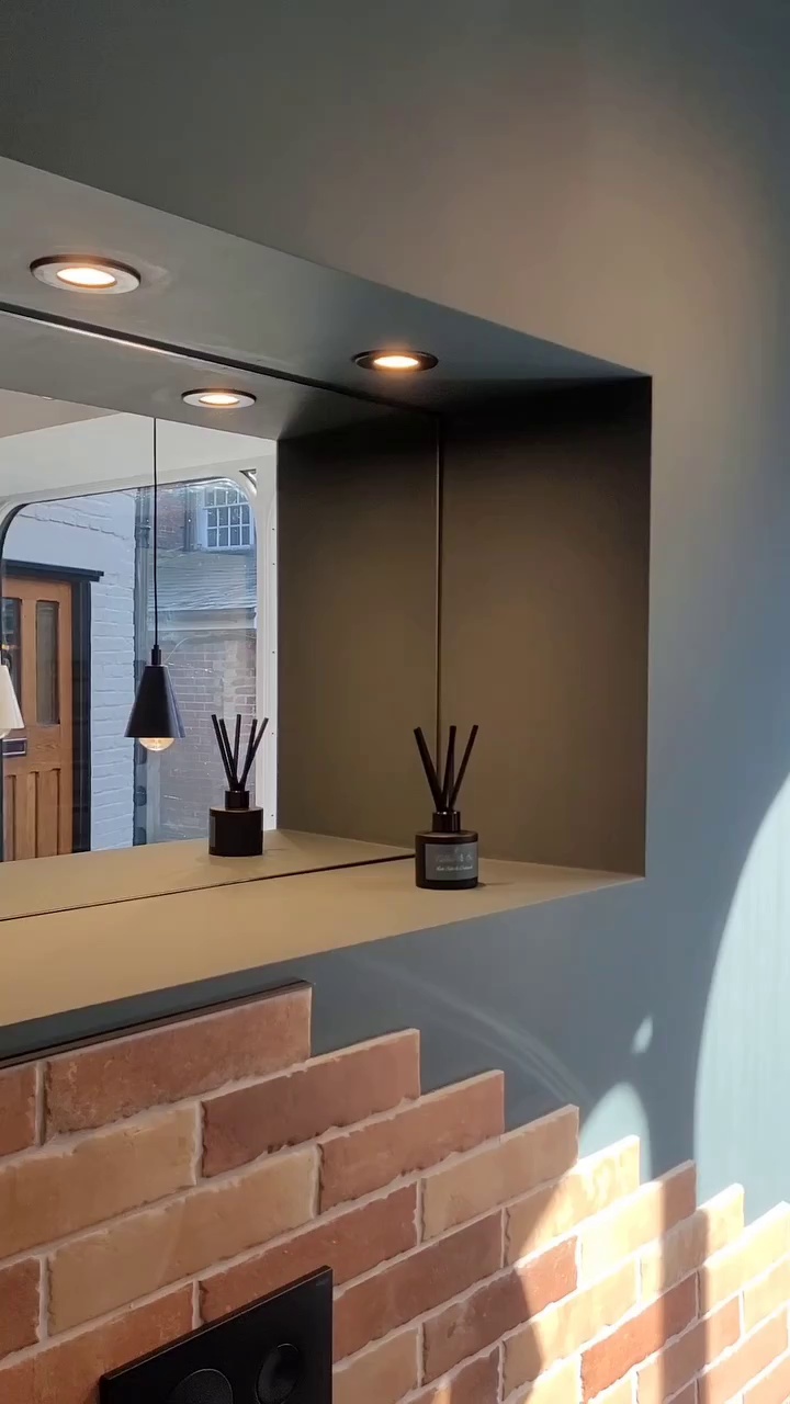 Bathroom mirror with built-in lights, black pendant light, and reed diffuser, above brick half-wall.