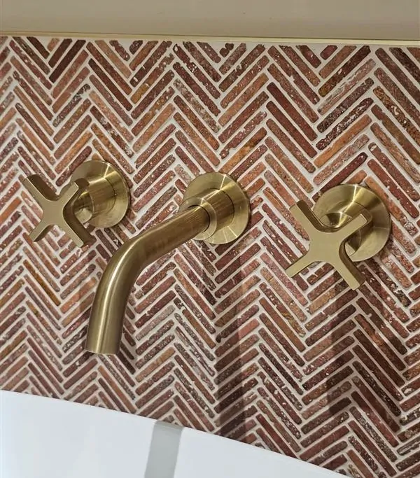 Brushed brass wall-mounted taps against decorative herringbone tiles