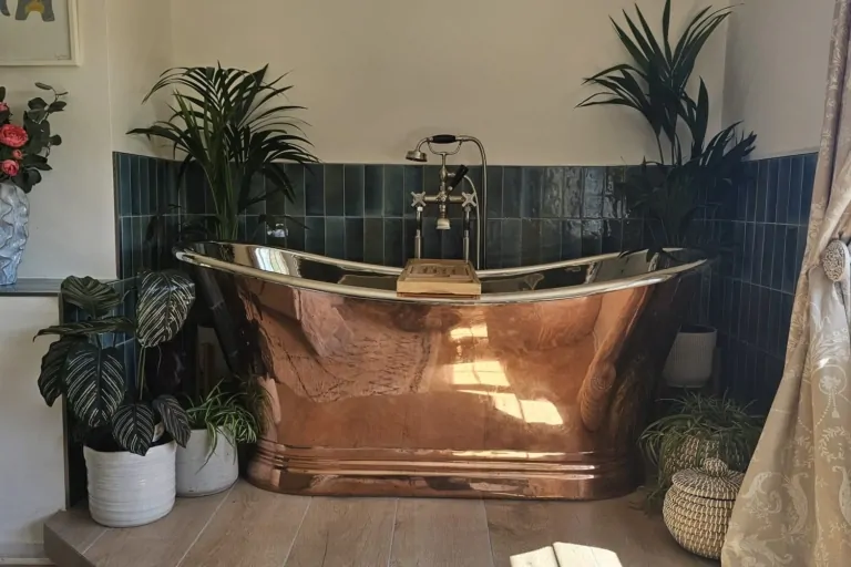 Copper and nickel freestanding bath surrounded by botanical plants and blue tiled walls