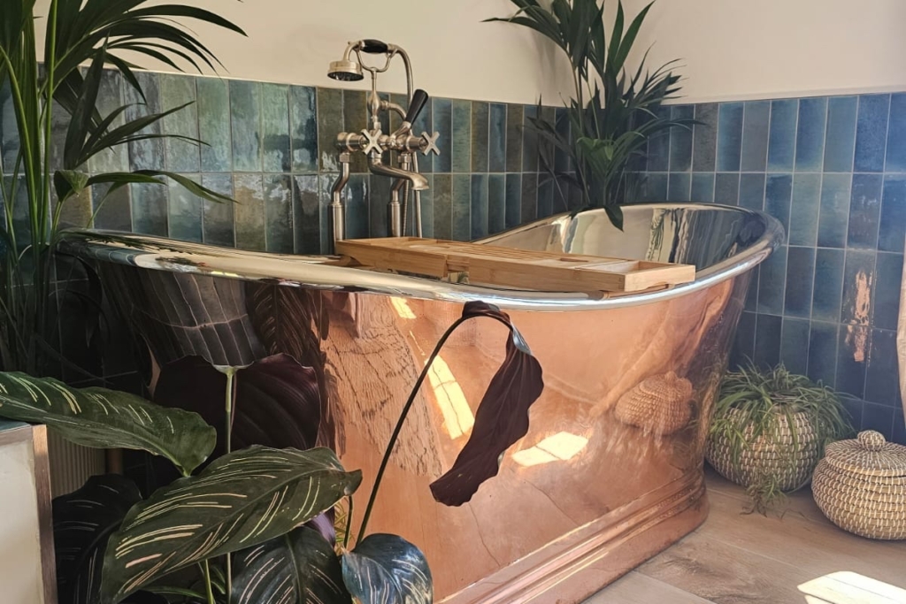 Copper and nickel freestanding bath framed by lush botanical planting and blue tiles