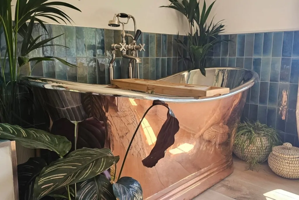 Copper and nickel freestanding bath framed by lush botanical planting and blue tiles